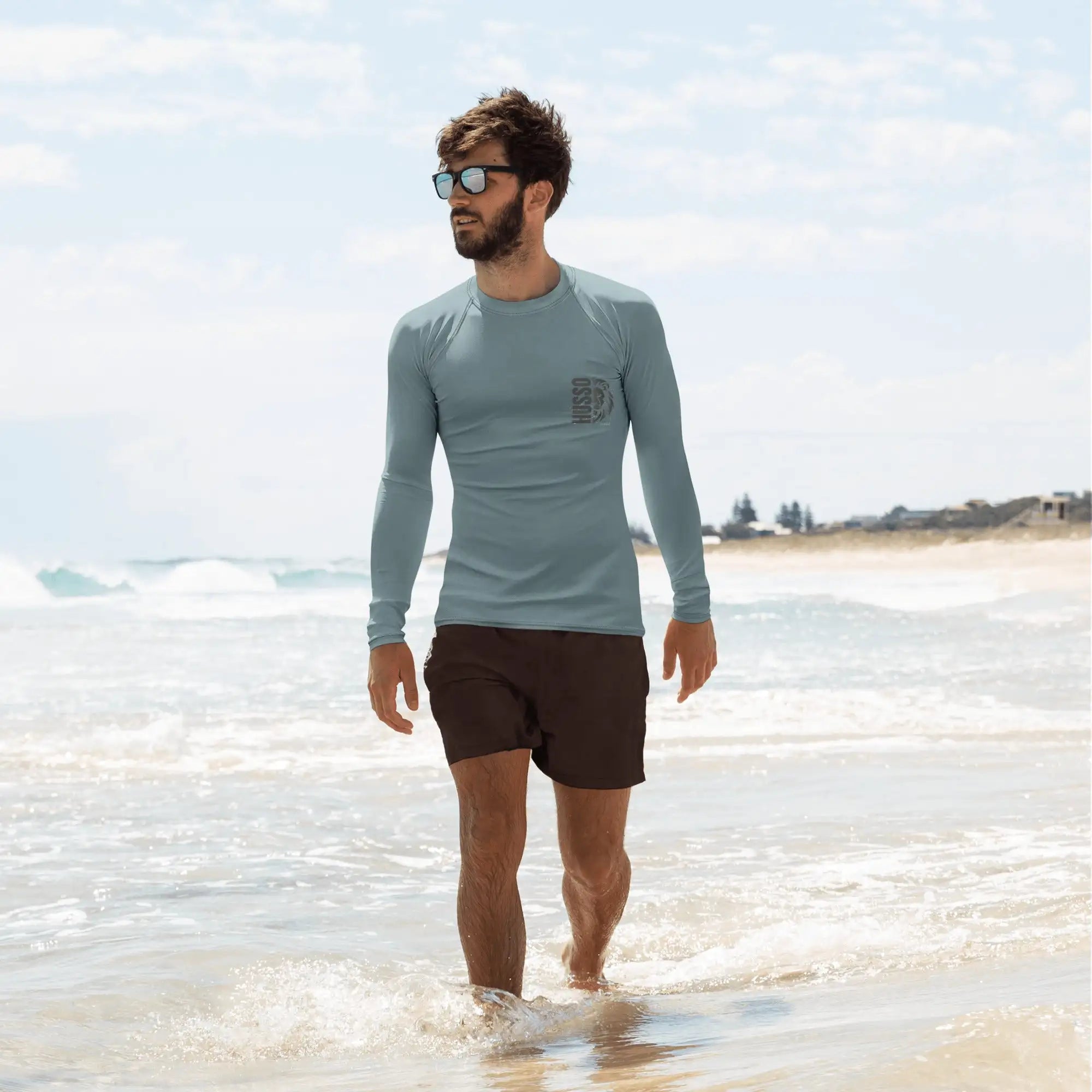 Man wearing teal Husso Hard long sleeve rash guard and black swim shorts walking on the beach