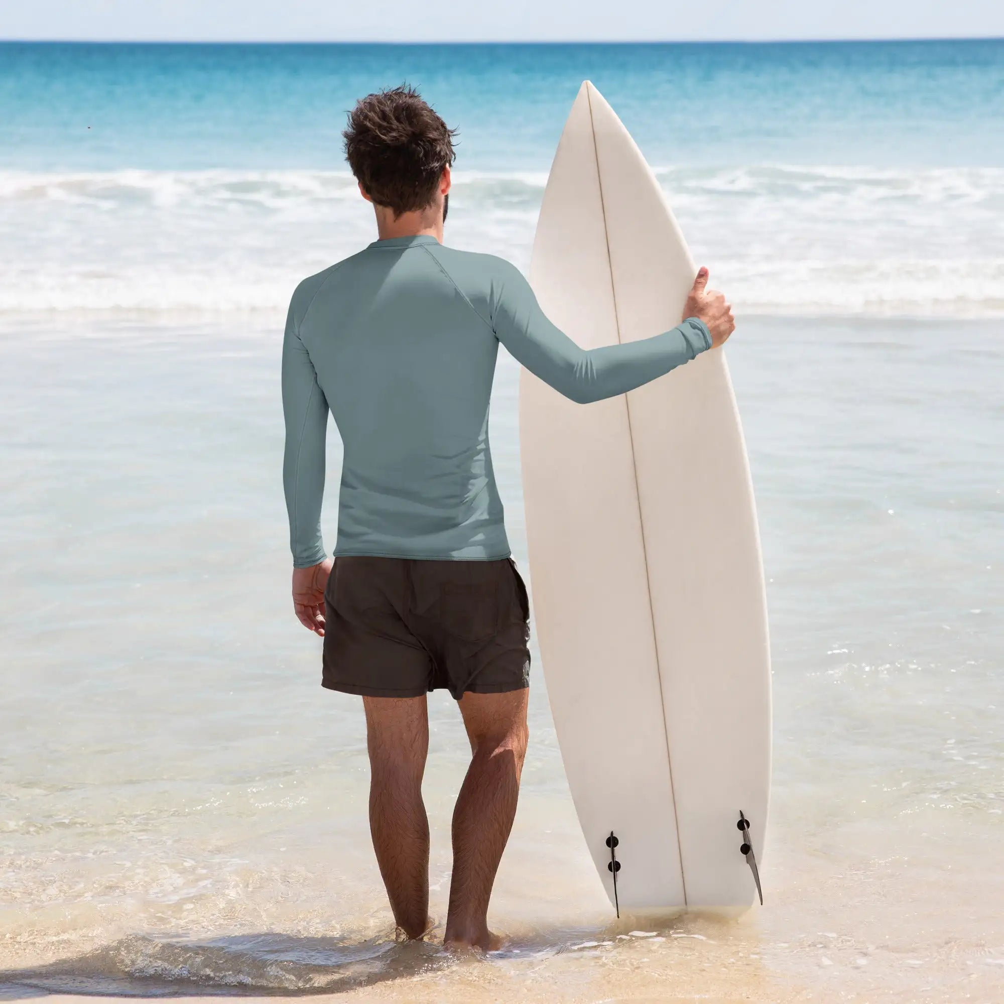 Back view of man wearing teal Husso Hard long sleeve rash guard holding surfboard at the beach